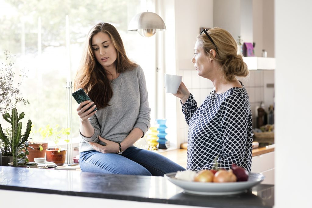 Jente med mobil, mor med kaffekopp på kjøkken