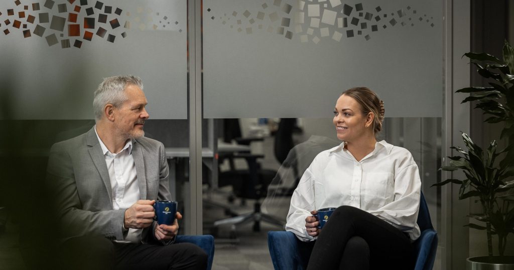 Mann i grå dessjakke med hvit skjorte sitter i en blå stol med en blå kaffekopp. mannen smiler bort til en dame med brunt hår og hviot skjorte som sitter i en blå stol ved siden av.