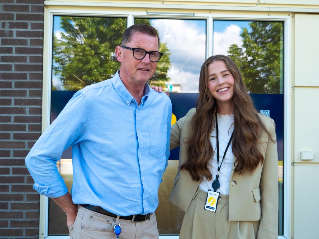 En mann i blå skjorte med briller smiler med en kvinne med langt brunt hår i beige klær. 