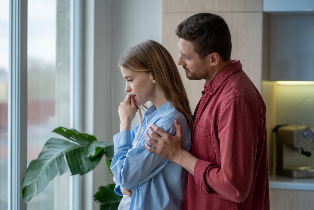 Ung kvinne med lyseblå skjorte står ved vinduet og er bekymret. En mann i rød skjorte holder rundt henne.