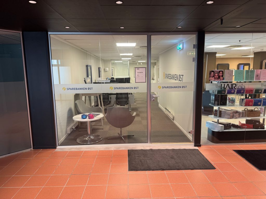 Kontoret er plassert i en gang med røde fliser på gulvet sammen med glassvindu og dør. Inne i kontoret er det hvite vegger med grått vegg-i-vegg teppe. To grå stoler står i hjørnet av lokalet ved glassvinduet. På glassvinduet og døren er det en hvit foliering med Sparebanken Øst sin logo.