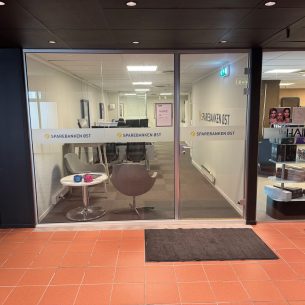 Kontoret er plassert i en gang med røde fliser på gulvet sammen med glassvindu og dør. Inne i kontoret er det hvite vegger med grått vegg-i-vegg teppe. To grå stoler står i hjørnet av lokalet ved glassvinduet. På glassvinduet og døren er det en hvit foliering med Sparebanken Øst sin logo.